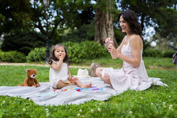 Cheerful female with toothy smile in striped casual dress with bubble blower having picnic with baby girl with teddy bear and crayons in park - Fotografie, Obrázek