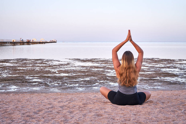 Gün batımında kumsalda yoga yapan uzun saçlı genç bir kadın. Kafkasyalı uzun sarı saçlı kız gün doğumunda kumlu bir sahilde yoga egzersizleri yapıyor..                   - Fotoğraf, Görsel