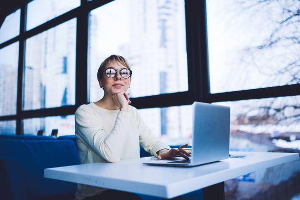 Glad female freelancer in casual clothes with eyeglasses looking at camera sitting at table with hand at chin using laptop while working alone - Fotó, kép