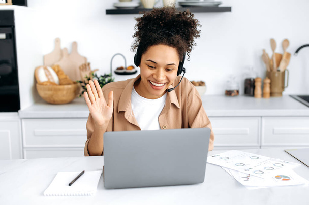 Çevrimiçi video bağlantısı. Pozitif başarılı Afro-Amerikan kıvırcık saçlı, kulaklıklı kız, evinde mutfakta oturuyor, bilgisayarını kullanarak iş arkadaşlarıyla konuşuyor. - Fotoğraf, Görsel