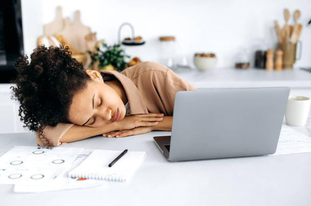 Privación crónica del sueño. Chica afroamericana agotada, freelancer o estudiante, se quedó dormida en el escritorio cerca de la computadora portátil, sobrecargada de trabajo o estudio en línea, necesita descansar y dormir - Foto, Imagen