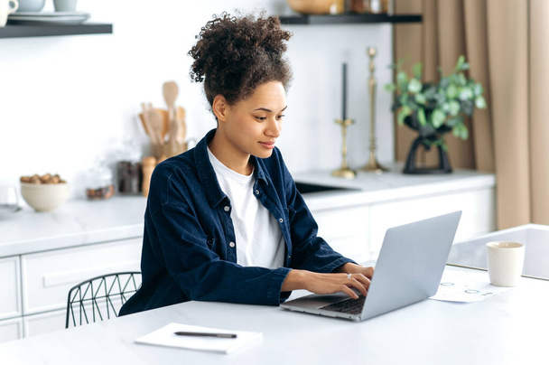 Focused smart pretty mixed race curly haired girl, designer, IT specialist, sitting at home in the kitchen, using laptop to work online from home, working on new project, strategizing, analyzing - Fotó, kép