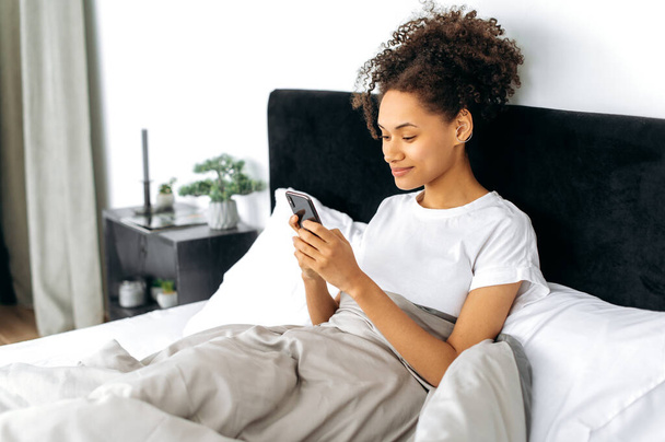 Szerkentyűk és technológiai koncepció. Elégedett nyugodt afro-amerikai lány, ül az ágyban reggel, használja az okostelefonját, beszélgetett a barátaival a közösségi oldalakon, reggeli híreket olvasott, válaszolt e-mailben - Fotó, kép