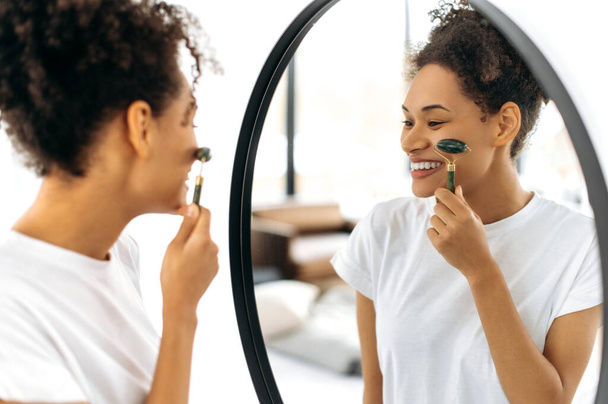 Афроамериканская красавица в белой футболке, стоит перед зеркалом дома, делает свой утренний номер красоты, заботится о своей коже лица, использует лицевой нефритовый ролик, предотвращает морщины, улыбается - Фото, изображение