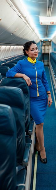 飛行機の中で助手席の近くに立っている間にカメラと笑顔を見ていると制服を着た楽しい女性スチュワーデス - 写真・画像