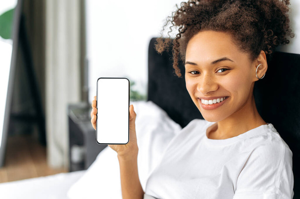 Close-up of pretty curly African American girl with freckles, sits in a bed, demonstrates her smartphone with a blank white mock-up screen for advertising or presentation, looks at camera, smiling - Foto, Imagem
