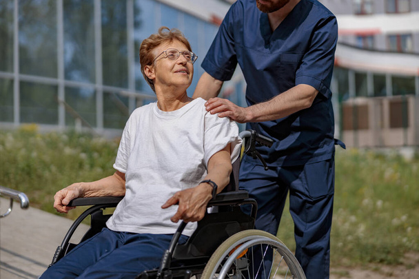 Nurse talking to patient on wheelchair in hospital yard . Rehabilitation concept. - Photo, Image