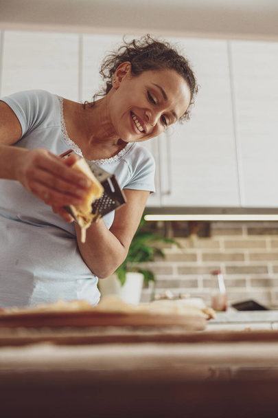 Nainen, jolla on hymy kasvoillaan, raastaa juustoa pizzan päälle ruoanlaitossa keittiössä. - Valokuva, kuva