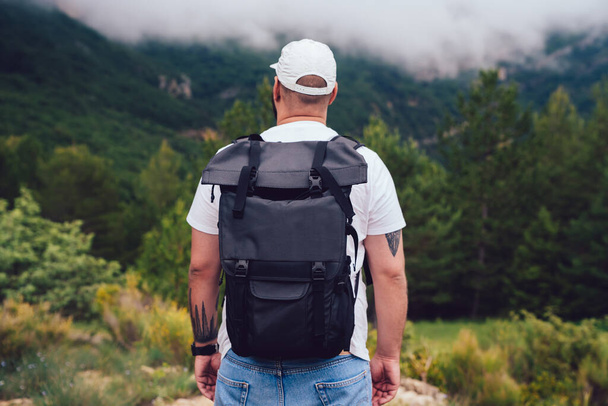 Back view of anonymous male tourist with backpack and hat standing on edge of steep rock while spending time in forest - Φωτογραφία, εικόνα