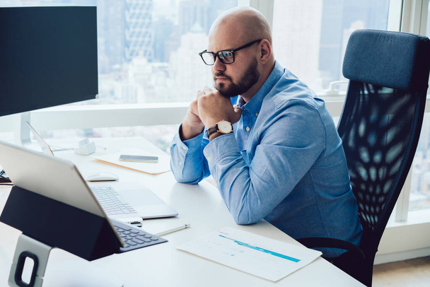 Oldalnézet a töprengő fiatal szakállas férfi vállalkozó olvasás információt laptopon, miközben ül az asztalnál támaszkodik a kezét koncentrált munka közben - Fotó, kép