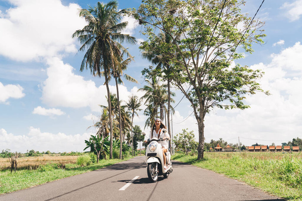 Joyful young lady with long dark hair in white shirt riding motorcycle on asphalt road though green meadows during summer vacation in tropical country - Zdjęcie, obraz