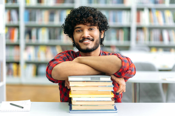 Positive clever indian or arabian guy, mixed race male, university student, sits at a desk with books, in the library against the background of bookshelves, looks at camera, smiles friendly - Zdjęcie, obraz