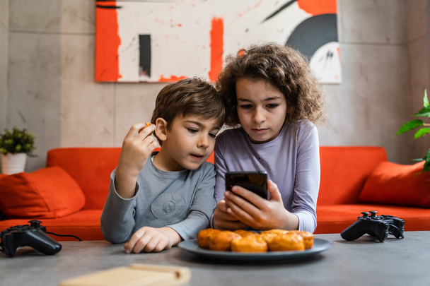 Siblings brother and sister boy and girl or children friends using mobile phone smartphone at home in room to watch video make a call or play online games leisure family concept real people copy space - Zdjęcie, obraz