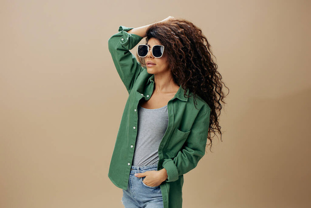 STYLE CONCEPT. Beautiful tanned curly Latin female in casual things sunglasses touch natural hair posing isolated over pastel beige background look aside. Copy space Mockup Banner. Fashion offer - Photo, Image