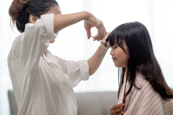 Mãe asiática cortando cabelo para sua filha na sala de estar em casa, enquanto ficar em casa a salvo de Covid-19 Coronavirus durante o confinamento. Conceito de auto-quarentena e distanciamento social
. - Foto, Imagem