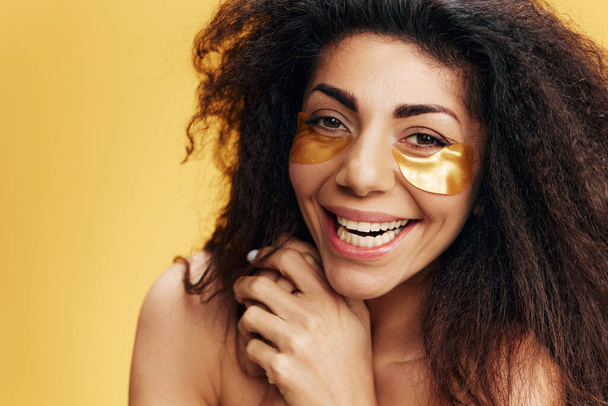 Onnellinen kihara kaunis latinalaisamerikkalainen nainen, jolla on kultaiset hydrogeelilaastarit silmän alla, nauraa kameralle, joka poseeraa keltaisella taustalla. Kosmeettinen tuote mainos Natural kauneus käsite Studio muotokuva - Valokuva, kuva