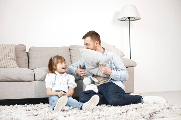 Loving father playing with his daughter with Down syndrome at home together. Man and girl sitting on a floor near sofa. Bearded man wearing blue shirt and girl white shirt. - Foto, immagini