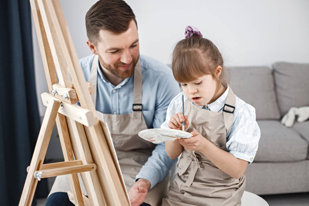 Little girl with Down syndrome wearing white shirt and beige apron. Girl with her father is painting on easel together. Father helping his daughter to paint. - Zdjęcie, obraz