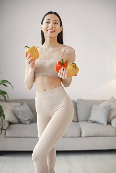 Slim and beautiful woman wearing beige leggins and top and holding fresh red and yellow paprika in hands. Young girl with fit body posing at home. Sport, fitness, diet, nutrition and health care - Fotó, kép