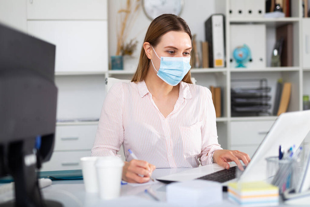 Businesswoman in protective medical mask is working with project behind laptop in the office - Valokuva, kuva