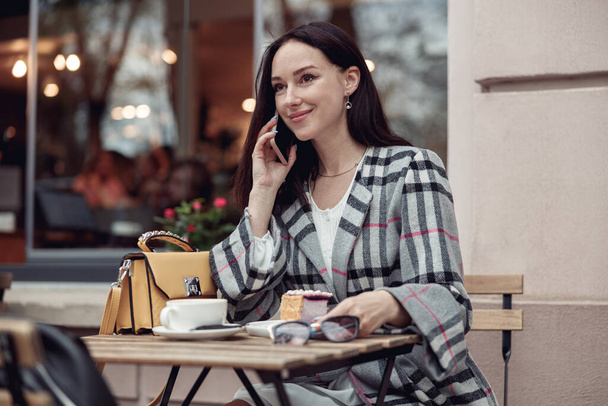 カフェでチーズケーキとコーヒーを飲みながら携帯電話で話す魅力的な若い女性。肖像画. - 写真・画像