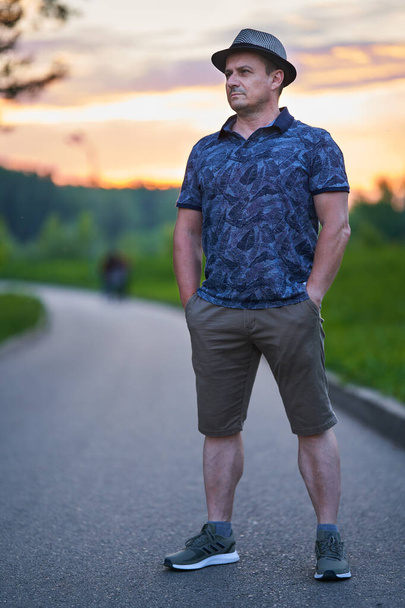 Portrait of a trendy Caucasian man in summer outfit with a hat in a park, urban environment, at sunset - Foto, imagen