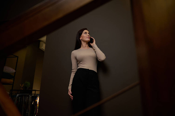 Confident attractive businesswoman, entrepreneur, designer in casual attire talking on mobile phone, gossiping standing on the stairs in the hallway of a modern interior design office. Bottom view - Fotografie, Obrázek