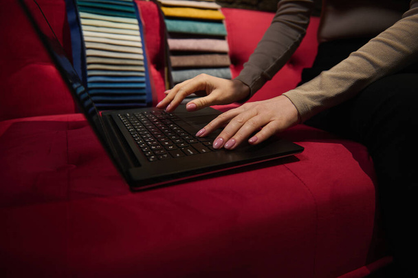 Подробности: Руки неузнаваемой женщины, читающей текст на клавиатуре ноутбука, сидящей на красном велюровом диване с цветной обивкой на заднем плане, планирующей новый проект дизайна интерьера дома - Фото, изображение