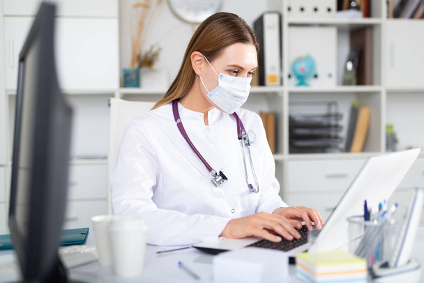 Portrait of young female doctor in face mask working on laptop in clinic office - Photo, Image