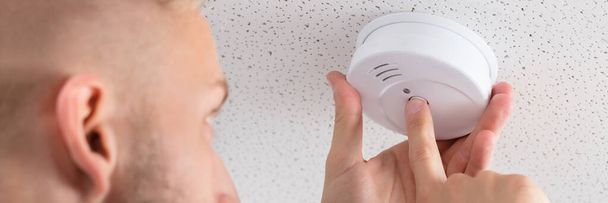 Low Angle View Of A Person's Hand Installing Smoke Detector On Ceiling Wall At Home - 写真・画像