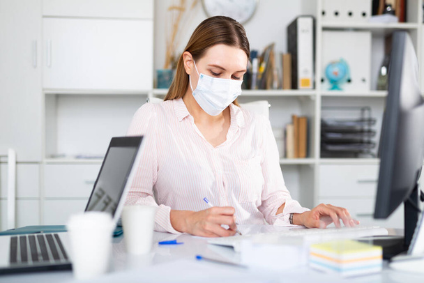 Businesswoman in protective mask working alone with laptop and papers in office, new normal due to coronavirus outbreak - Photo, Image