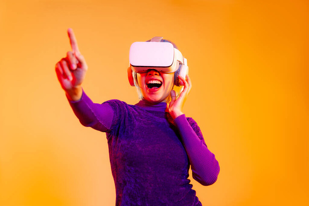 Metaverse concept happy woman in purple shirt wearing vr goggles headset finger pointing on the orange screen background colorful neon like cyberpunk.	 - Foto, immagini