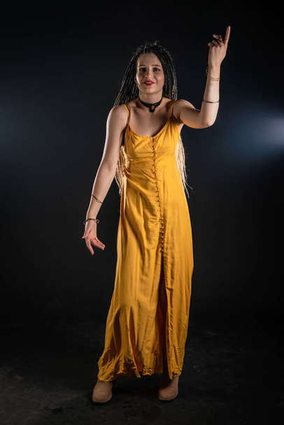 Mystic αρχαία θεά βασίλισσα φορώντας ένα κίτρινο φόρεμα σε ένα απομονωμένο μαύρο φόντο στούντιο. Γυναίκα φεμινίστρια που φοράει κοτσίδες και ποζάρει σε στούντιο. - Φωτογραφία, εικόνα