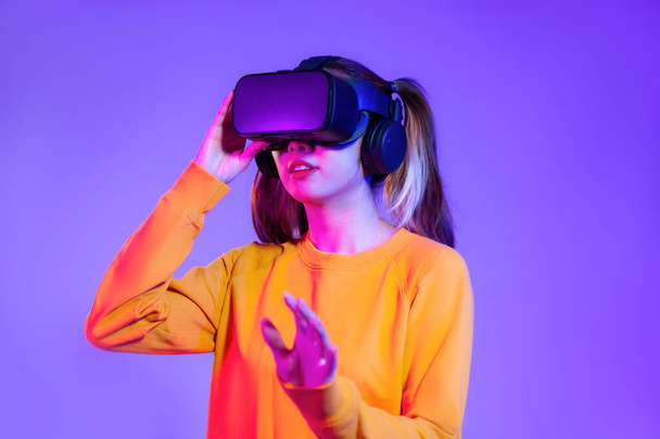 Young asian woman in orange sweatshirt wearing black vr headset watching playing and hand touching on the purple background. Metaverse concept vr future girl lifestyles - Fotó, kép