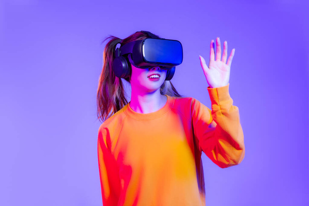Young asian woman in orange sweatshirt wearing black vr headset watching playing and hand touching on purple background. Metaverse concept vr future girl lifestyle. - Foto, immagini