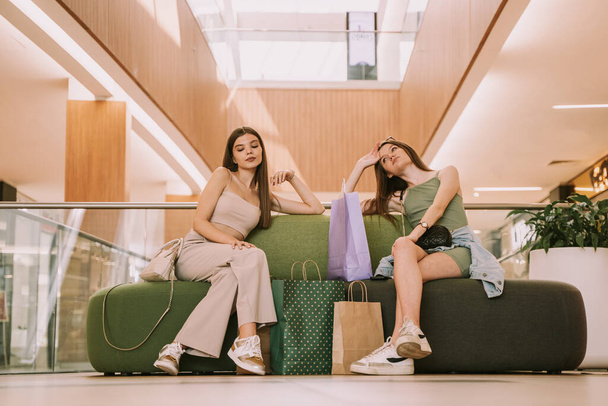 Two young and beautiful friends sitting on a couch at the mall exhausted and finally ready to go home - Foto, imagen