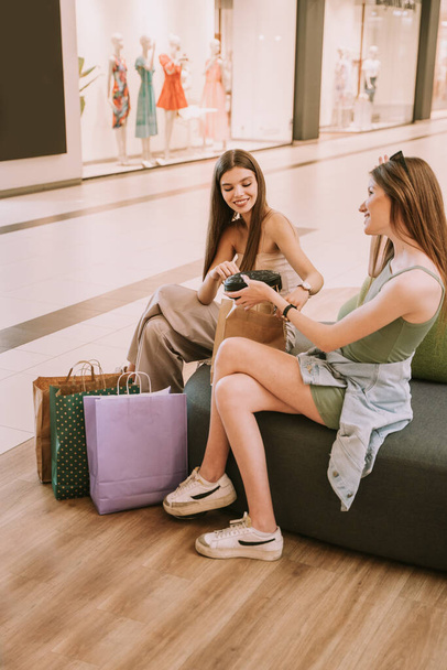 Alışveriş merkezindeki kanepede oturan iki güzel arkadaştan biri diğerine çantayı gösteriyor. - Fotoğraf, Görsel