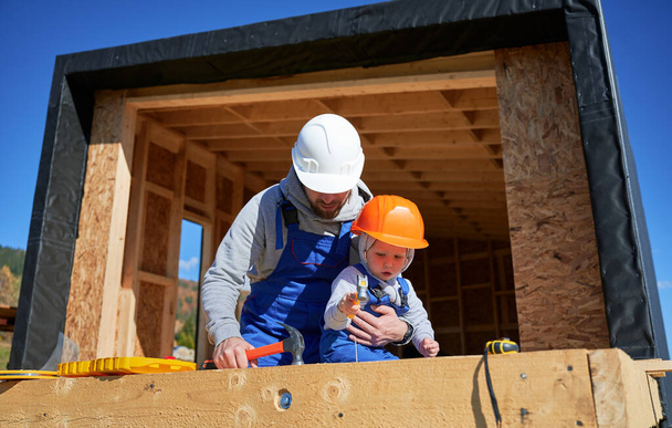 Father with toddler son building wooden frame house. Male builders hammering nail into plank on construction site, wearing helmet and blue overalls on sunny day. Carpentry and family concept. - Фото, изображение