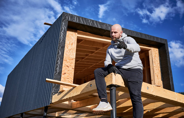 Άντρας προγραμματιστής κτίριο ξύλινο σπίτι πλαίσιο για τα θεμέλια σωρό. Πορτρέτο του φαλακρού άνδρα που κάθεται στη βεράντα στο εργοτάξιο, επιθεωρώντας την ποιότητα της εργασίας. - Φωτογραφία, εικόνα
