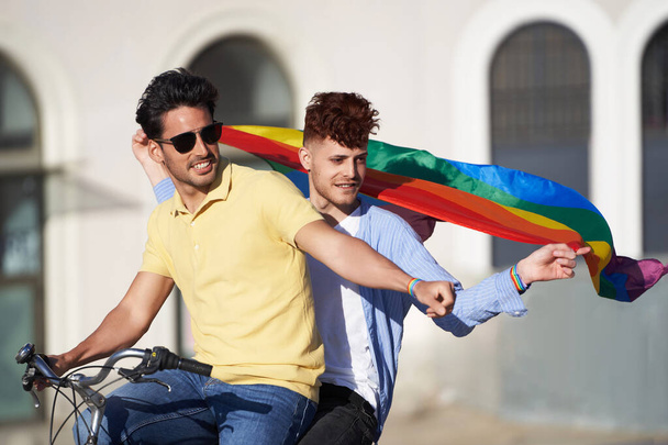 Молодая пара друзей с флагом гей-гордости во время езды на велосипеде на открытом воздухе. Концепция ЛГБТ, отношений и равноправия. - Фото, изображение