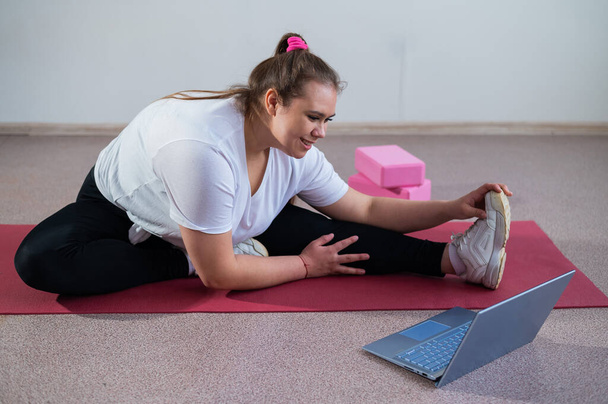 Young caucasian fat woman doing bends on a sports mat and watching a training video on a laptop. A chubby girl doing stretching remotely using video communication. - Zdjęcie, obraz