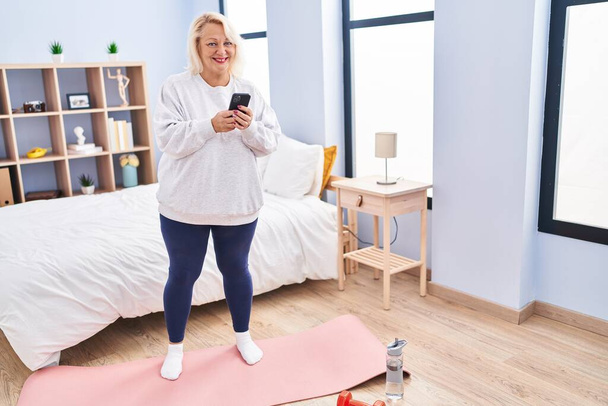 Middle age blonde woman using smartphone standing at bedroom - Foto, Bild