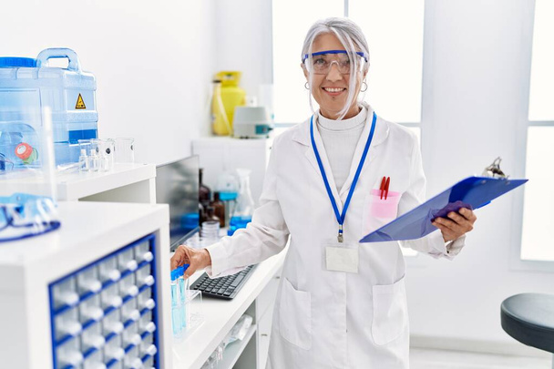 Седовласая женщина средних лет в научной форме держит планшет и пробирку в лаборатории - Фото, изображение
