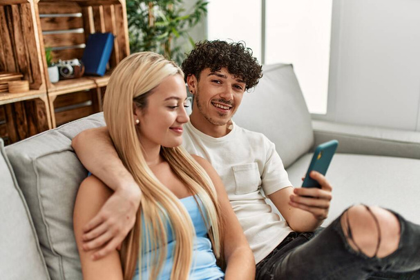 Молодая пара с помощью смартфона сидит дома на диване. - Фото, изображение