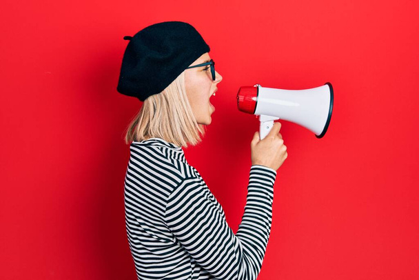 Caucasian blonde woman wearing glasses and beret screaming with megaphone over red background - Photo, Image