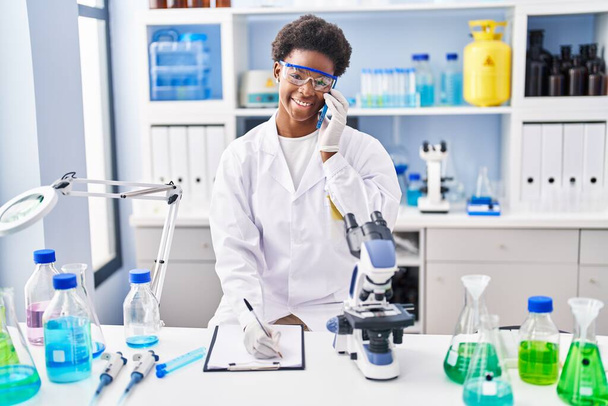 Αφροαμερικανή γυναίκα φορώντας στολή επιστήμονα μιλώντας στο smartphone γράφουν στο πρόχειρο στο εργαστήριο - Φωτογραφία, εικόνα