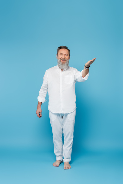 full length of bearded barefoot man in white clothes pointing with hand on blue  - Фото, изображение