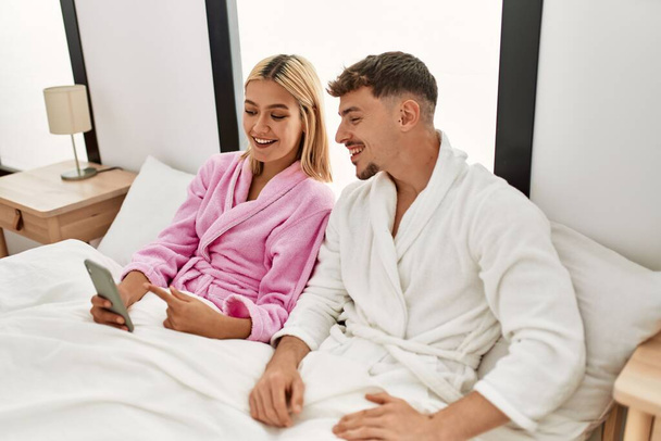 młoda kaukaska para uśmiechnięta szczęśliwy za pomocą smartfona siedzi na łóżku w domu. - Zdjęcie, obraz