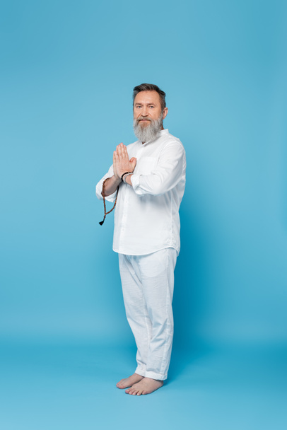 bearded spiritual mentor with rosary standing with praying hands on blue background - Photo, Image
