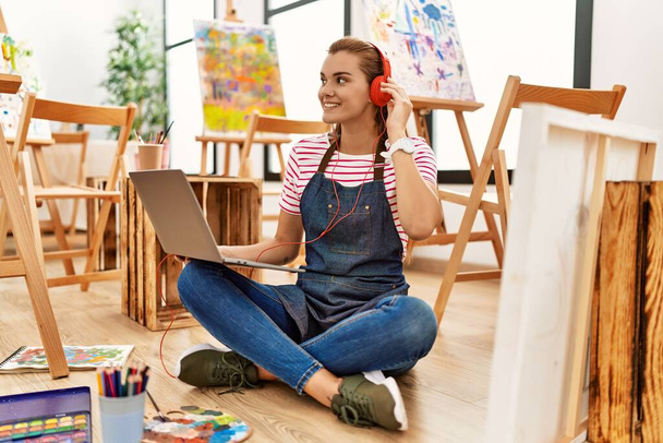 Giovane donna sorridente fiducioso utilizzando laptop e cuffie in studio d'arte - Foto, immagini